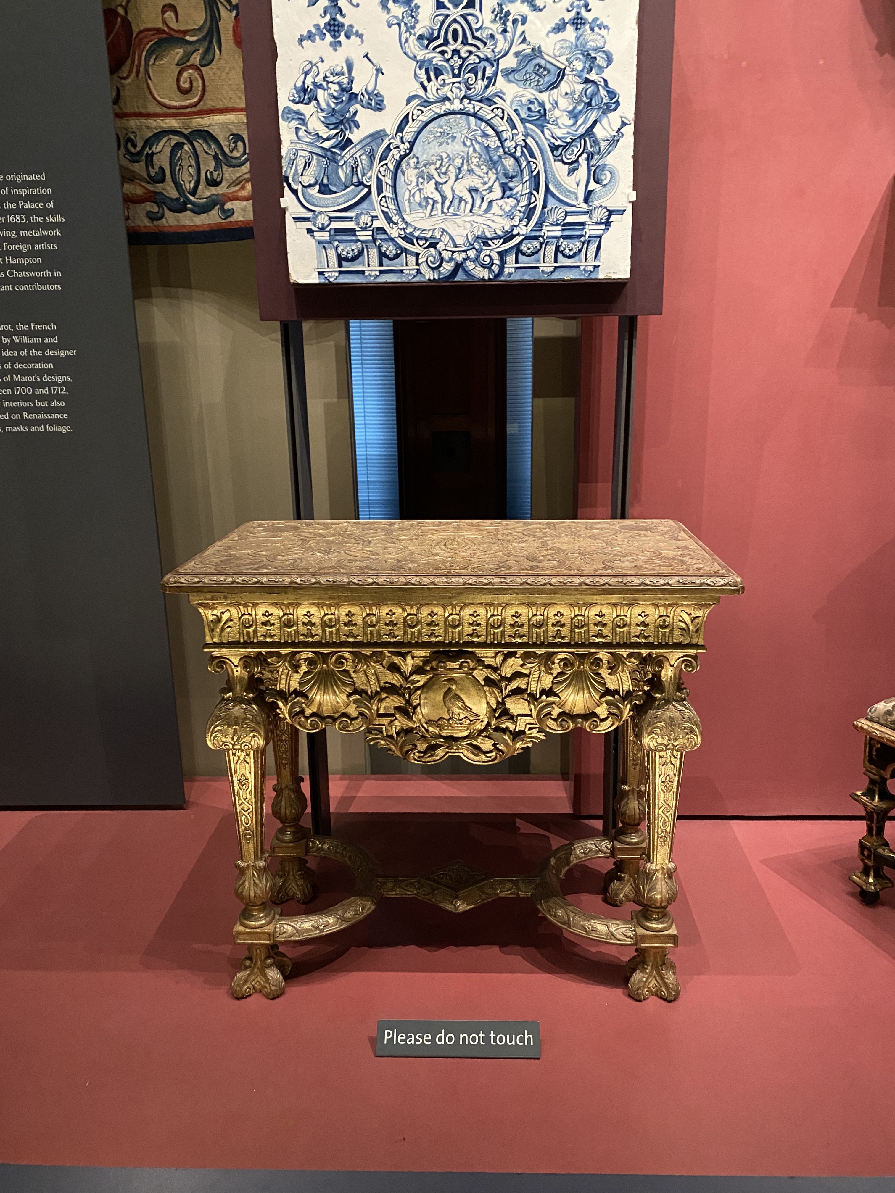 the image shows a gilded, ornate antique table with a marble top, displayed in a museum setting. above the table, there's a decorative blue and white tile panel. a small sign on the floor reads "please do not touch".