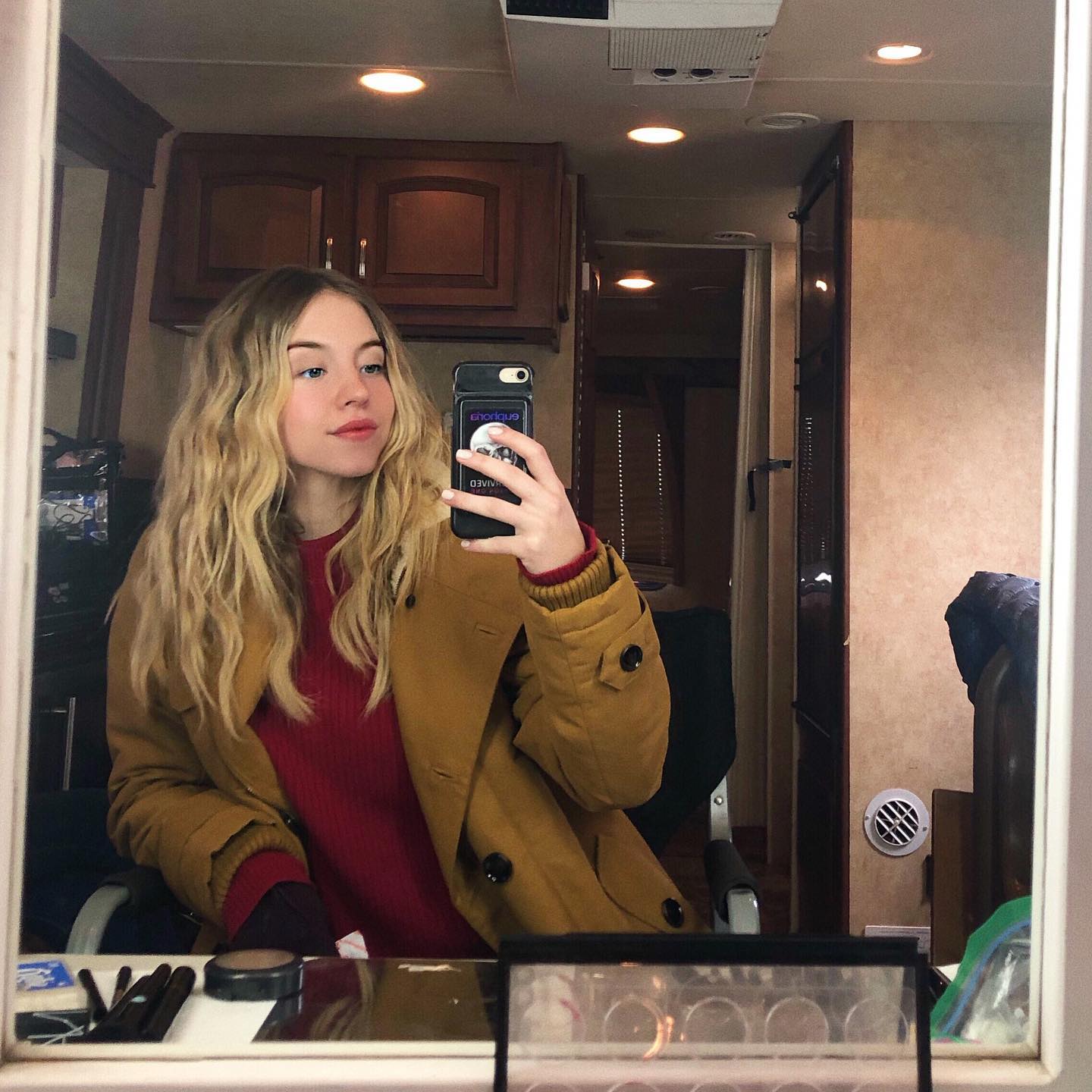 a young blonde woman wearing a mustardcolored coat and a red sweater takes a selfie in a mirror. the mirror reflects a makeup vanity and the interior of what appears to be a film or television production trailer.  she holds her phone up to take the picture, and various makeup items are visible on the vanity in front of her. the overall setting suggests a behindthescenes glimpse of a film or television production.