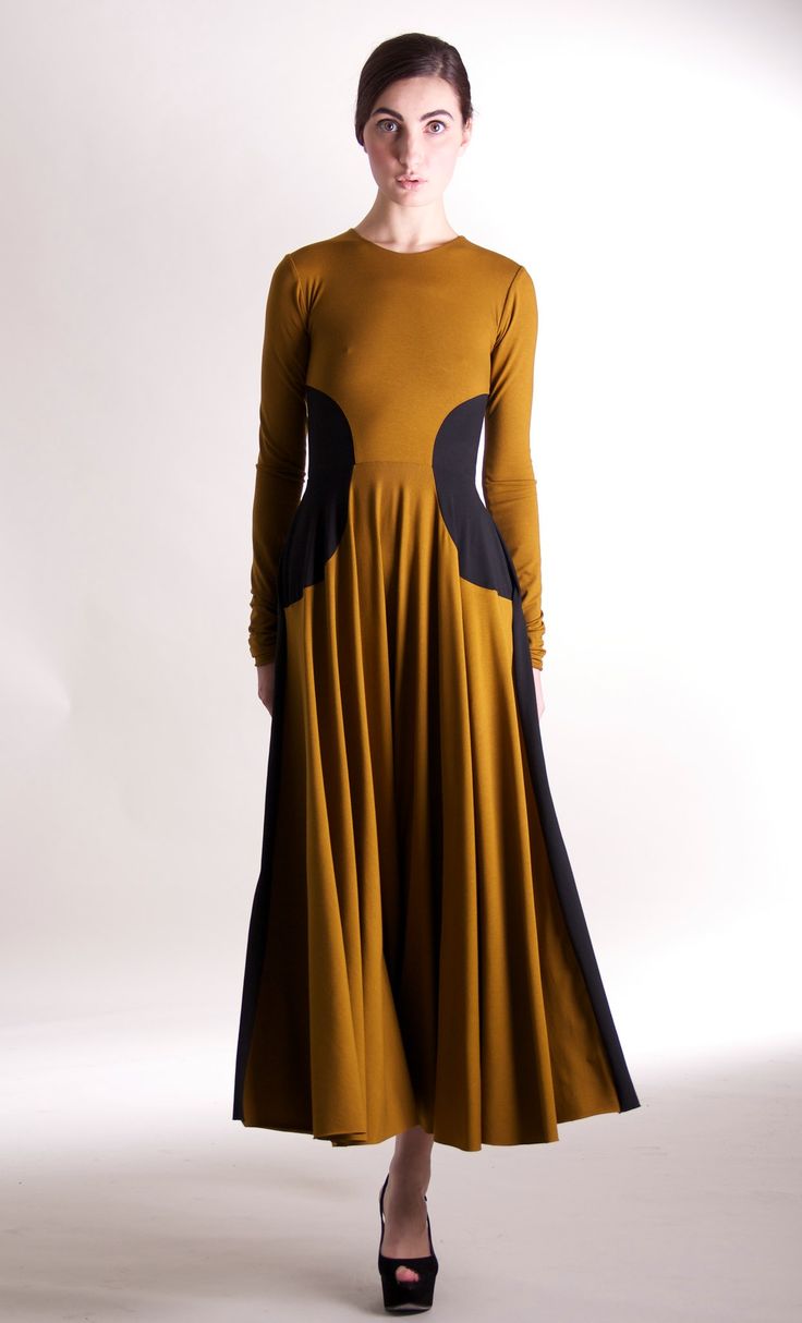 a young woman in a mustard yellow dress with black cutouts around the waist stands in a studio. the dress is a long skirt with a slightly fitted top and long sleeves. she has long, straight dark hair and she's wearing black highheeled shoes. she's looking at the camera with a slightly surprised expression on her face.  she's posing with her hands down at her sides.