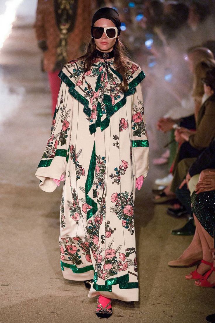 a model walks down a runway wearing a long, creamcolored floral dress with green sequin trim. the dress has wide sleeves and a high collar.  she's accessorized with sunglasses, a black headband, and pink shoes with embellishments. the background shows other attendees of a fashion show, slightly blurred. the overall aesthetic is high fashion and elegant.