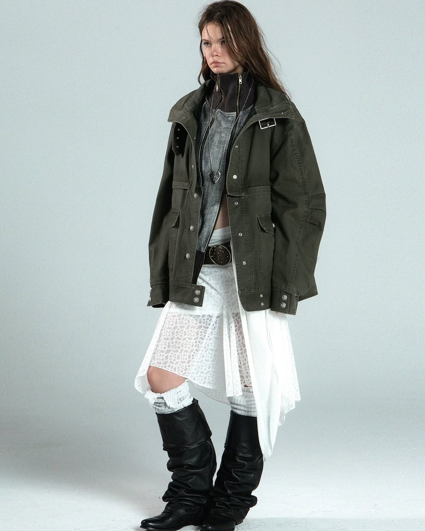 a fulllength studio shot of a young woman modeling a stylish outfit. she's wearing an oversized olive green militarystyle parka over a layered ensemble featuring a grey top and a white lace and pleated asymmetrical skirt.  she's accessorized with a necklace and belt and is wearing tall, slouchy black boots. the overall style is a blend of casual, grunge, and street style elements. the background is a plain light grey, focusing attention on the clothing and model.