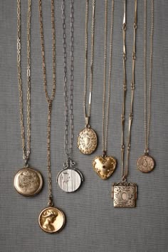 a close up photo of 8 necklaces with lockets hanging from them, on a grey fabric background.  the lockets are all different shapes and sizes and have various designs. the chains are all different sizes and styles. some of the chains are gold, some are silver, and some are a combination of the two. the lockets are all made of metal and have a variety of finishes, including gold, silver, and antique.  the necklaces are arranged in a way that showcases the different styles and designs.  the background is a soft grey which provides a nice contrast to the necklaces and lockets.  this is a beautiful and elegant image that would be perfect for a fashion magazine or website.