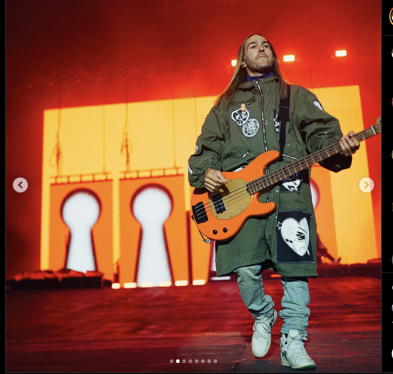 a musician in a green jacket and white pants plays a bright orange bass guitar on a stage with three large white keyhole shapes on the backdrop.  they are looking off to the right with their long hair flowing over their shoulders.