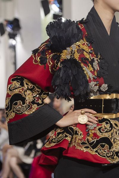 closeup shot showcasing the intricate details of a haute couture garment. the dress is primarily red with elaborate gold and black embroidery, featuring a unique design that incorporates cloudlike patterns and other symbolic motifs.  a large, black feather embellishment is prominently placed on the shoulder.  the model's hand is visible, adorned with a large gold ring. the overall style is opulent and dramatic. the background is blurred, suggesting a runway or fashion show setting.