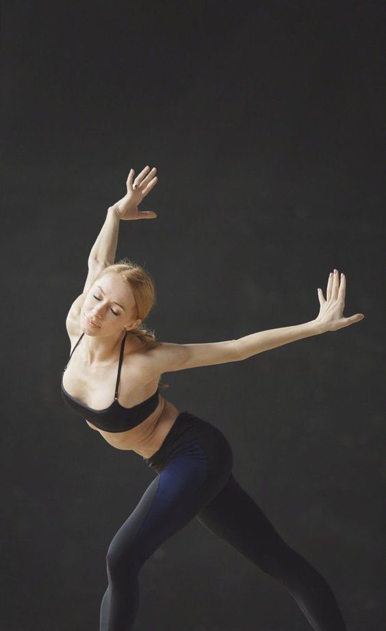 a woman in black workout wear executes a graceful dance pose against a black studio background.  her pose suggests flexibility and strength. the soft lighting emphasizes the contours of her body and creates a sense of movement.