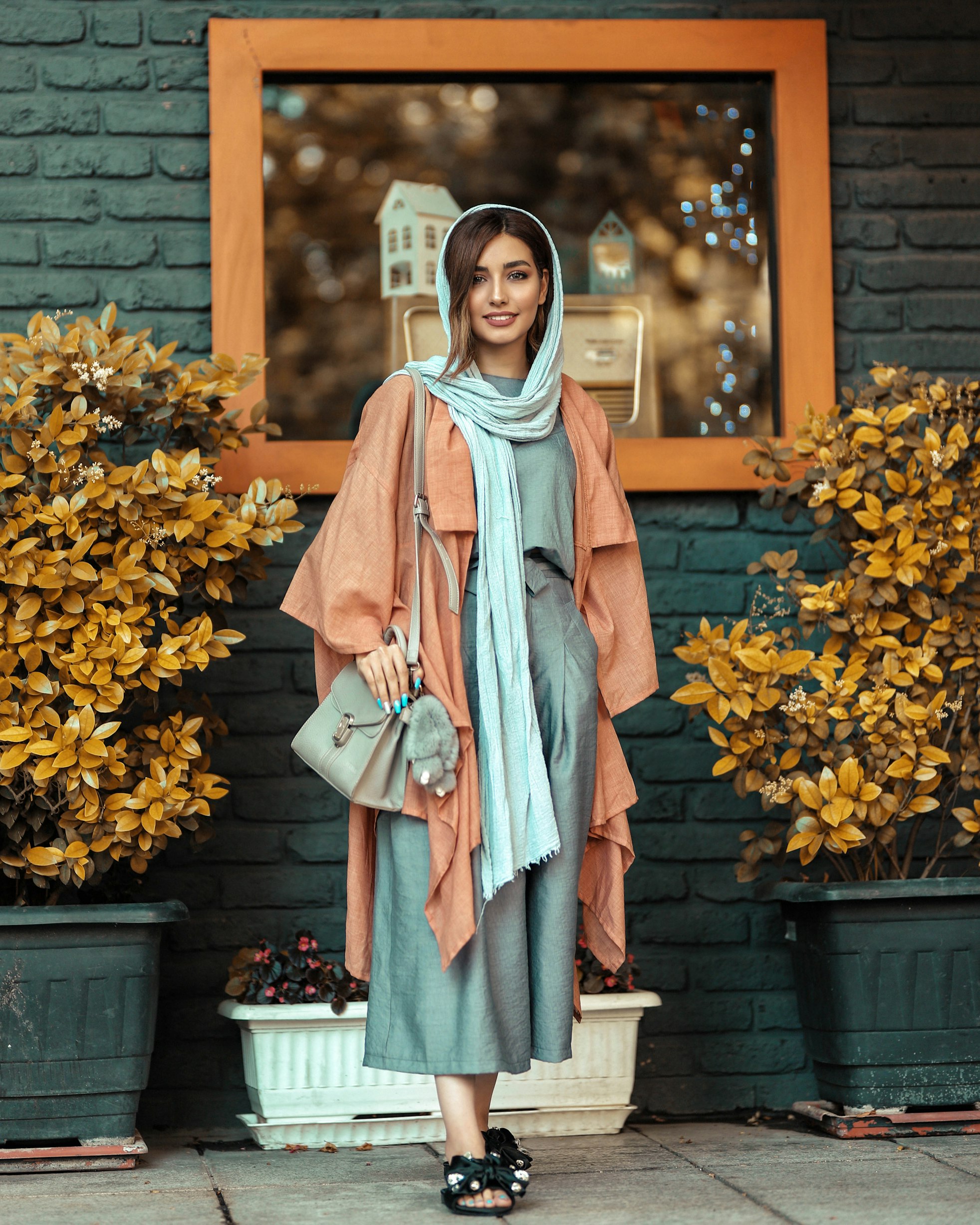 the image shows a stylish iranian woman standing in front of a brick building. she's wearing a peachcolored coat over a grey culottes, and a light blue hijab. she carries a lightgrey handbag. the overall style is modest yet fashionable. she is standing in front of a shop with a window displaying vintage items.  plants flank her on either side. the image has a soft, slightly vintage aesthetic.