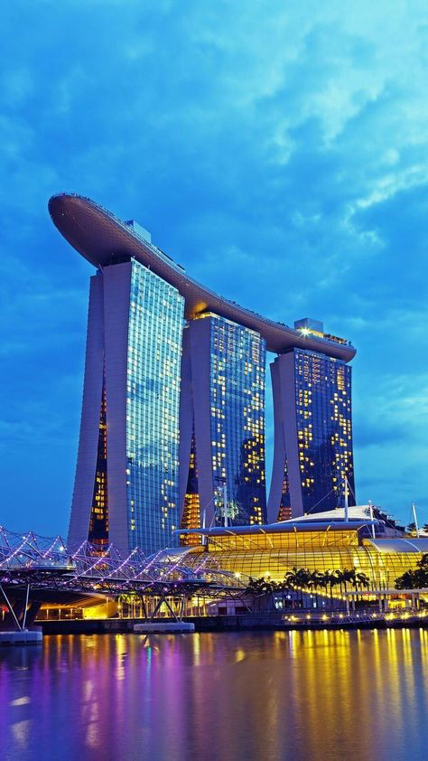 the image showcases the iconic marina bay sands resort in singapore at twilight.  the three towers of the hotel are brilliantly illuminated against a vibrant blue and purple sky.  the distinctive boatshaped structure on top is clearly visible.  the helix bridge is partially visible in the foreground, connecting to the hotel complex. the water reflects the lights of the city and the hotel, creating a stunning reflection. palm trees add a tropical touch to the scene, and a large glass structure, likely part of the hotel complex, is also prominent. the overall ambiance is one of modern luxury and vibrant city life at night.