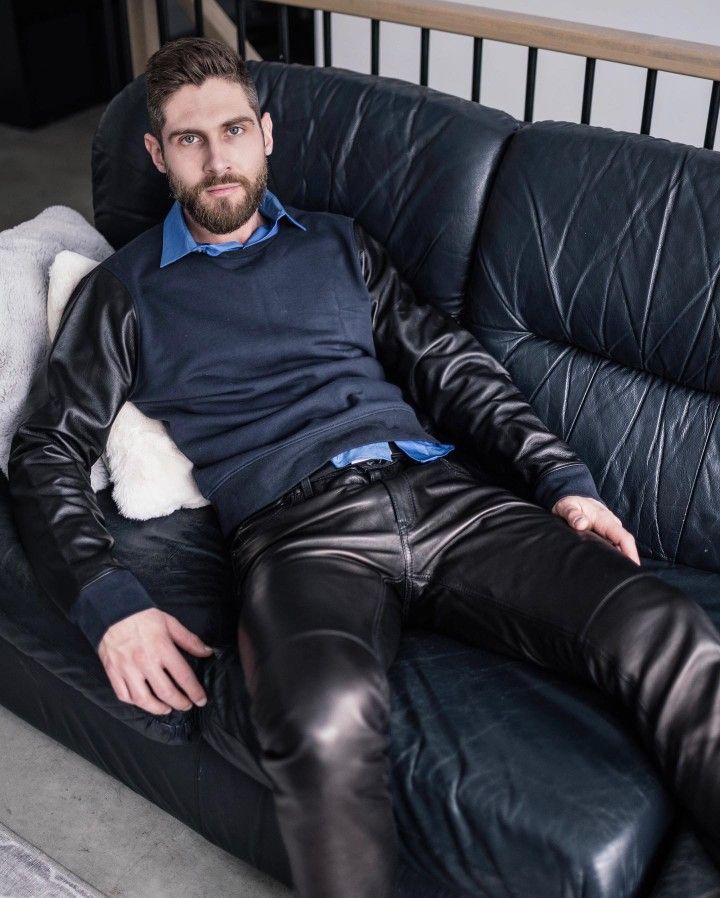 a man with a beard is sitting on a black leather couch. he is wearing a dark blue sweater with black leather sleeves, a blue collared shirt underneath, and black leather pants. he looks directly at the camera. the setting is indoors.