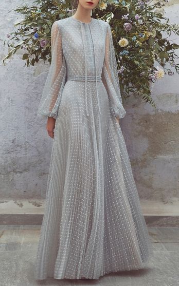 the image showcases a stunning, floorlength light blue dress. the dress is made of a pleated tulle fabric with delicate white polka dots.  it features long sheer sleeves with a slightly puffed shoulder detail and a high neckline with a subtle vshape. a fitted bodice flows into a full, pleated skirt. the dress is cinched at the waist with a matching fabric tie.  the model is posing against a backdrop of lush greenery and flowers, enhancing the dress's romantic and ethereal quality.  the overall aesthetic suggests a bridal or special occasion gown.