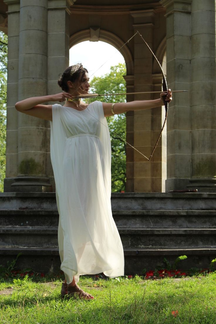 a woman dressed in a flowing white grecianstyle chiton or stola stands in front of a classicalstyle stone structure with columns. she's holding a bow and arrow, aiming with a focused expression, suggesting a portrayal of a greek goddess or archer. the setting appears to be an outdoor space, possibly the ruins of a temple, with sunlight illuminating her and the surrounding grassy area. she wears a gold headband and bracelets, adding to the classical aesthetic. the overall image evokes a sense of historical reenactment or theatrical costume photography.