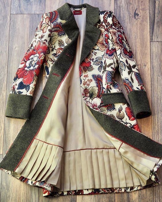 the image shows an open coat with a floral pattern on the exterior and a contrasting lining with pleats at the bottom. the coat has a dark green collar, cuffs, and lapel. the lining is a light beige color and the pleats add a decorative touch to the coat's interior. the floral pattern on the exterior features red, white, and blue flowers and leaves.