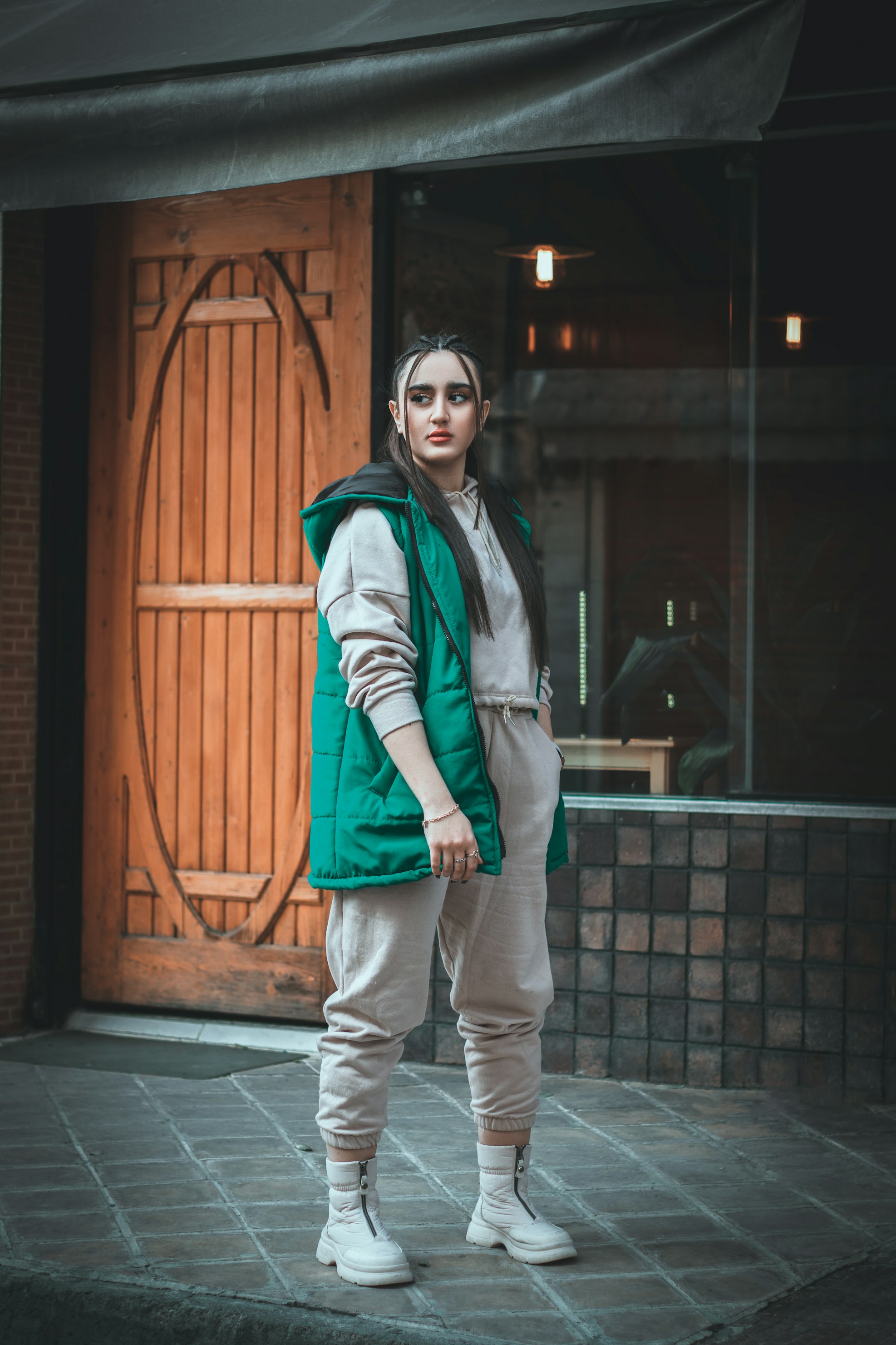 a young woman stands on a city sidewalk in front of a building with a wooden door and large window. she is wearing a teal puffer vest over a light beige sweatshirt and sweatpants, and white ankle boots. her dark hair is pulled back, and she has subtle makeup.  she appears confident and casual, looking away from the camera. the overall aesthetic is urban and stylish.