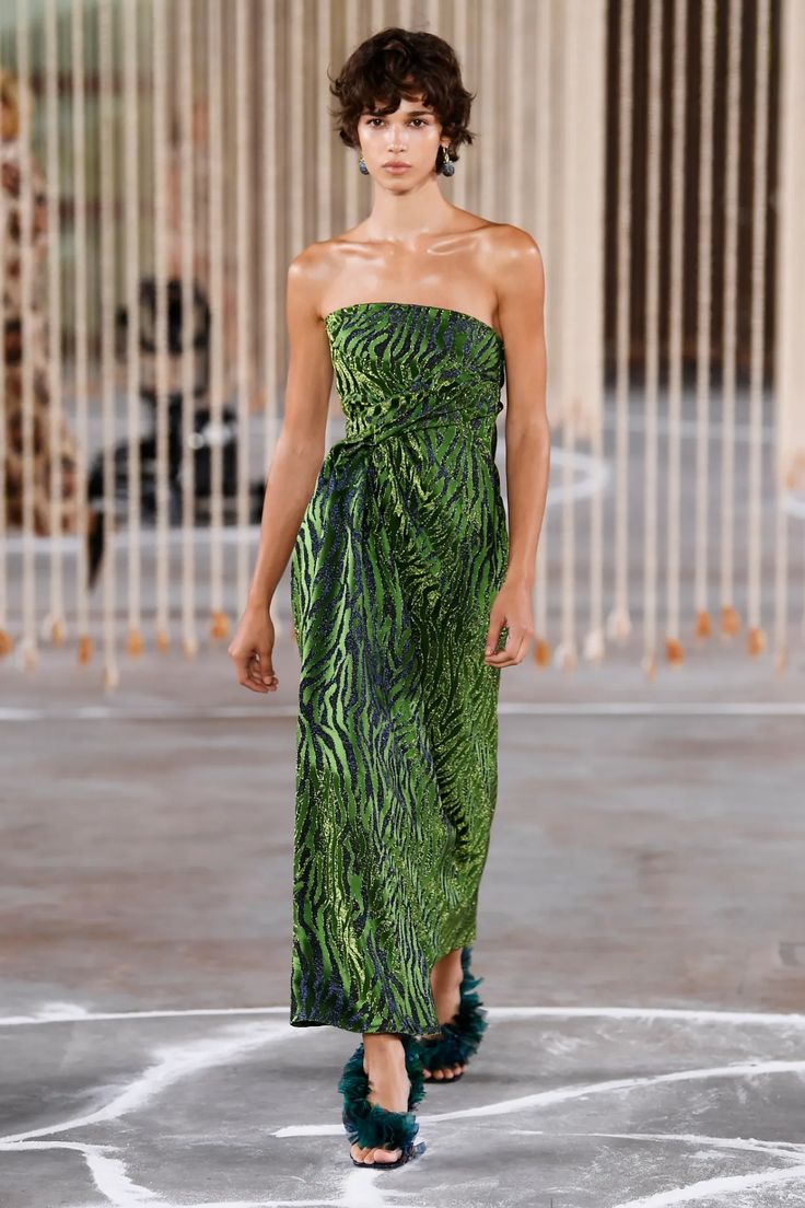 a female fashion model walks the runway wearing a vibrant green zebraprint maxi dress. the dress is strapless with a knotted waist detail. she is wearing dark green feathered heeled sandals and has short, curly hair. the background features a blurry setting with vertical lines suggesting a runway or studio backdrop. the overall mood is fashionable and stylish, showcasing the model and the dress in a professional fashion show context.