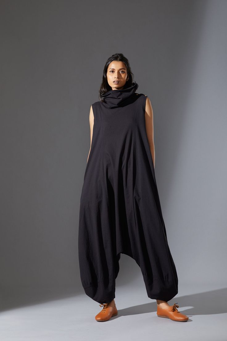 a woman wearing a black sleeveless jumpsuit with a high neck and baggy pants. she is wearing brown shoes and standing in a studio with a gray background. she is looking directly at the camera.