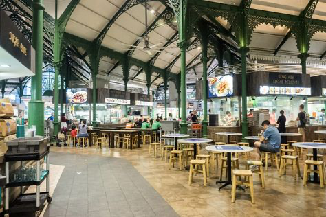 the image showcases the interior of a bustling hawker centre in singapore, possibly lau pa sat or telok ayer market. the space is characterized by its high, green metalframed ceiling and numerous wooden tables and stools arranged throughout.  various food stalls line the walls, each displaying their offerings. people are seated at the tables, enjoying meals. the atmosphere is lively and energetic, typical of a busy singaporean food court. the overall aesthetic blends traditional architecture with a modern, clean presentation of the food stalls.