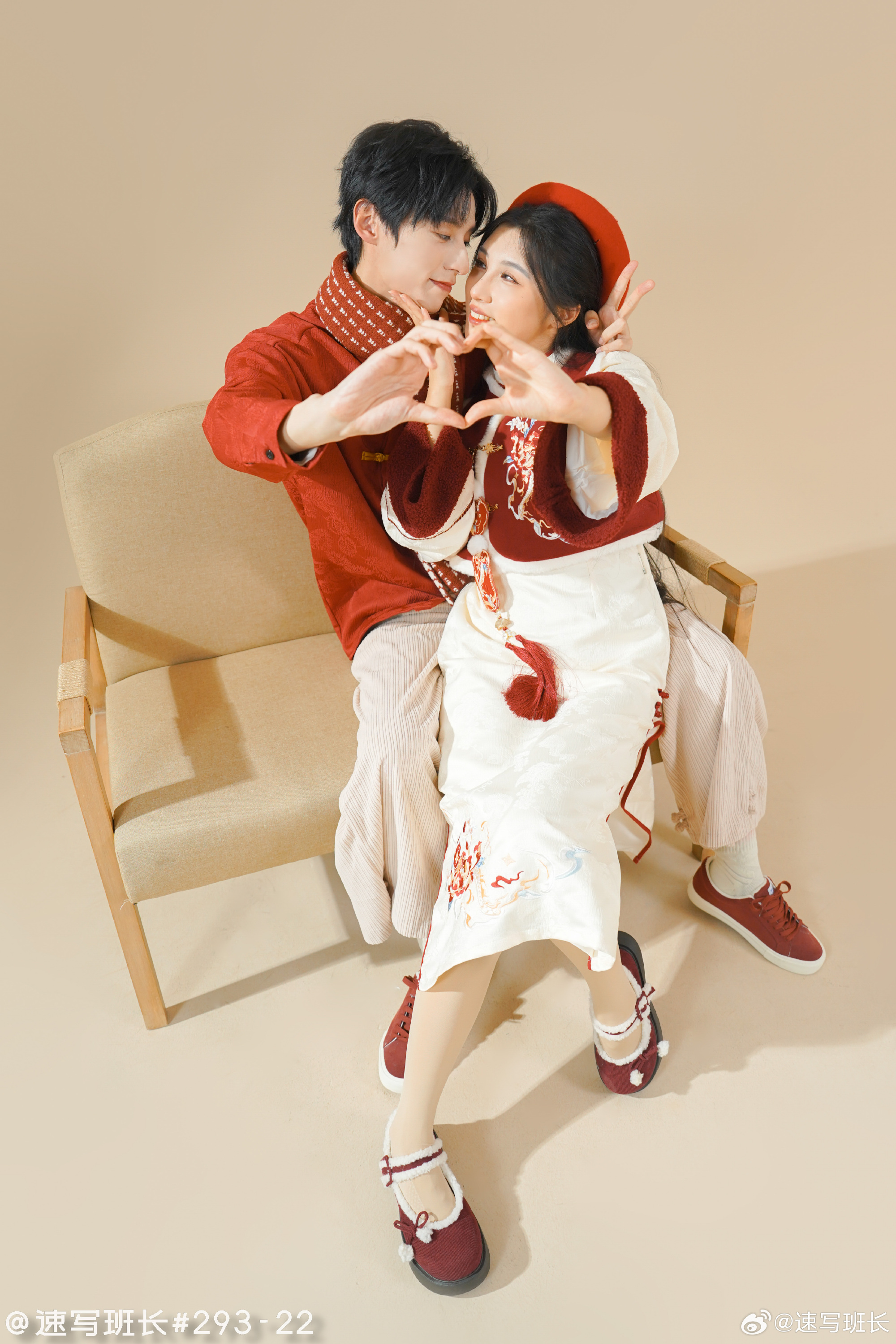 a young couple wearing traditional chinese clothing sit on a wooden chair in a studio setting.  the man is wearing a red shirt and scarf while the woman is wearing a white dress with red embroidery. they are both making heart shapes with their hands.