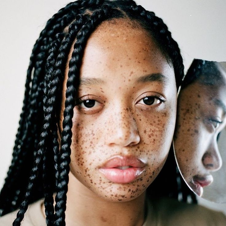 a closeup portrait of a young woman with freckles and braids. she is looking directly at the camera with a serious expression. a broken mirror is positioned next to her face, reflecting a profile view of her. the background is a neutral color.