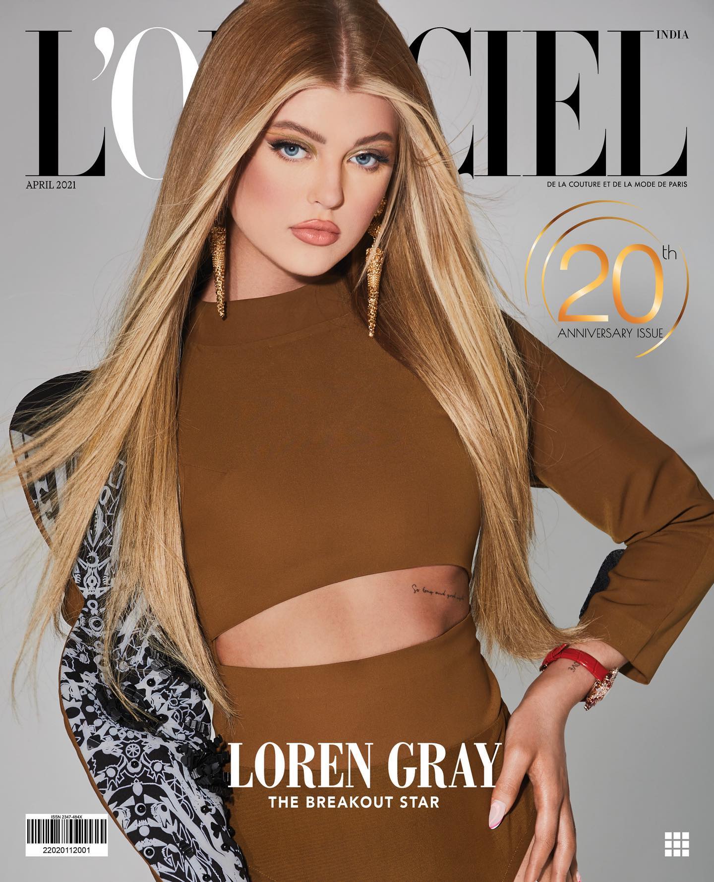this is the cover of l'officiel india magazine, april 2021 issue, featuring loren gray as "the breakout star". it is the magazine's 20th anniversary issue. loren gray is wearing a brown cutout dress and large gold earrings. she has blonde hair and blue eyes. there is a tattoo visible on her side. the cover also includes the magazine's name, date, and a barcode.