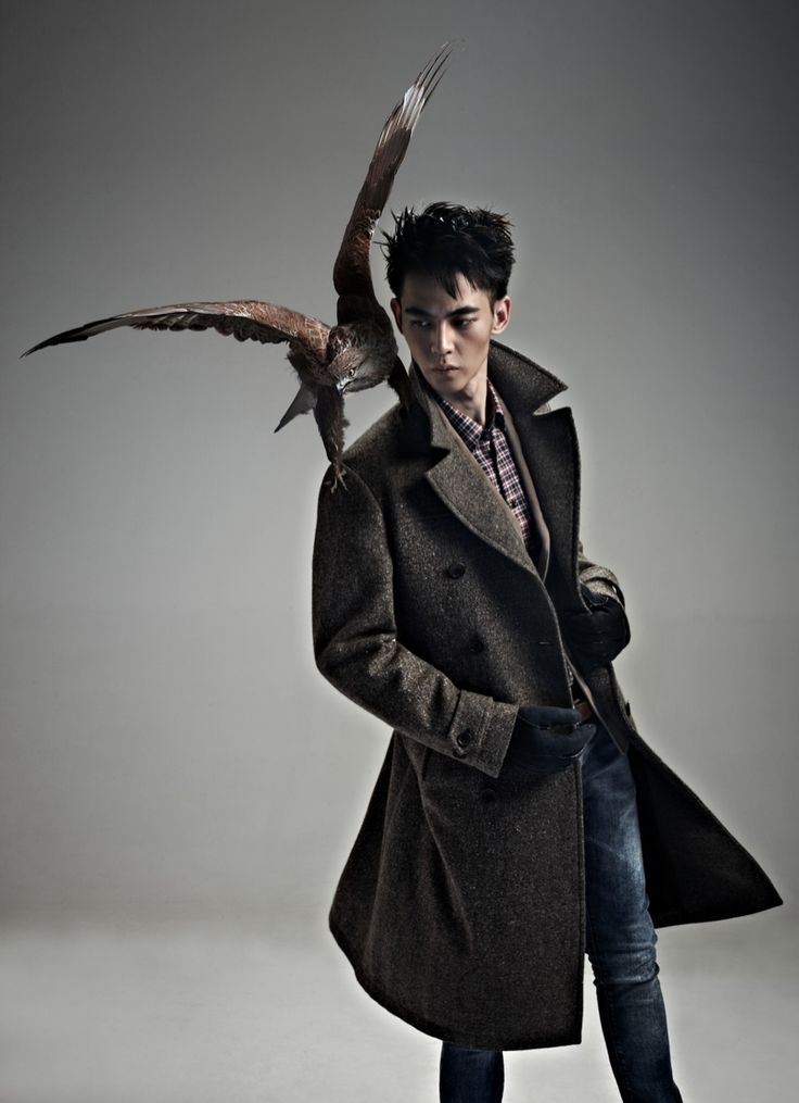 a stylish man wearing a black coat stands in a studio with a hawk on his shoulder. the man is looking away from the camera, and the hawk is looking at the camera. the background is gray.