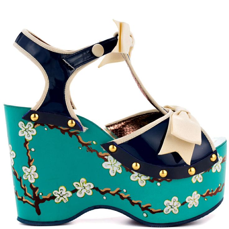 a closeup shot of a women's shoe, specifically a wedge heel platform sandal with a tstrap design. the sandal is primarily blue and teal with a floral pattern featuring white blossoms on a brown branch. a beige bow is attached to the front of the shoe. the shoe has gold studs along the upper edges. the style is bright and decorative.