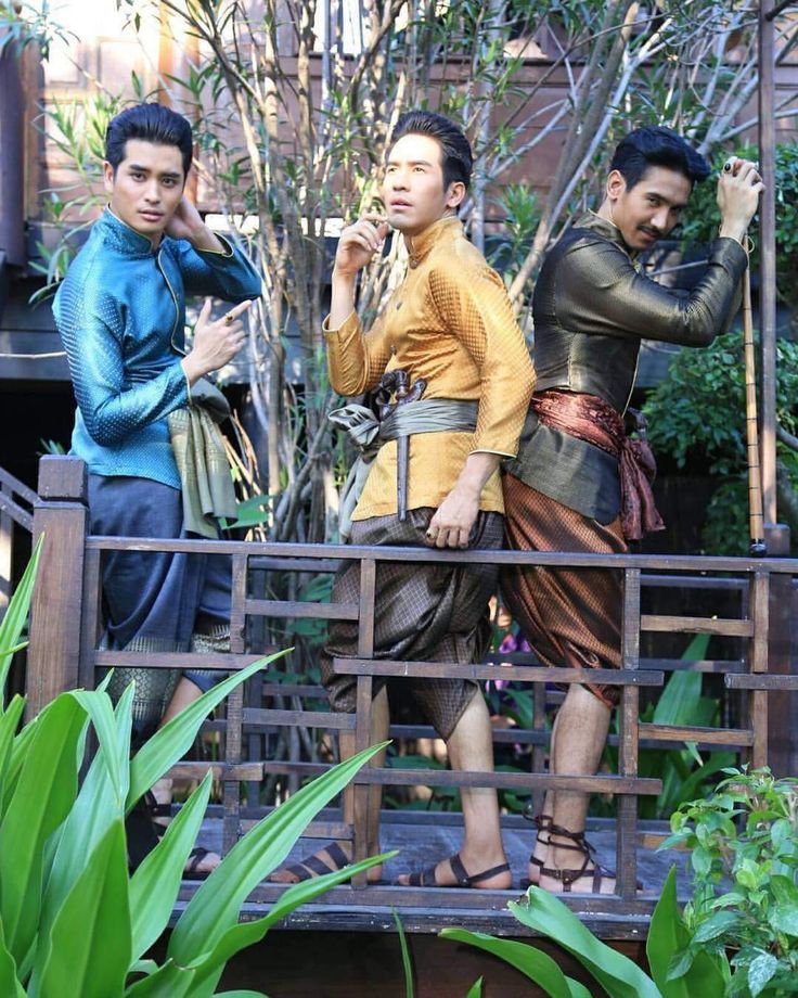three asian men are standing on a wooden platform with a railing. the men are wearing traditional thai clothing. two are smiling and one is laughing. the men are standing in front of a large green bush.
