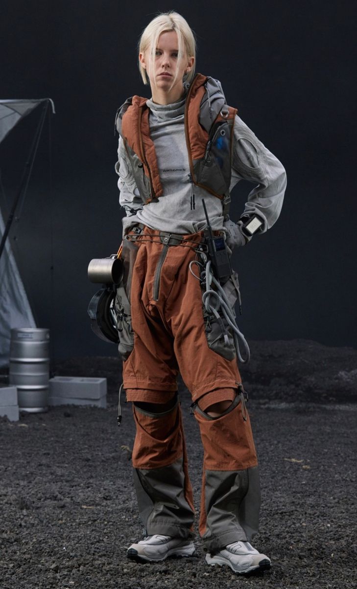 a full shot of a blonde female model standing on a dark, textured surface. she is wearing a layered outfit featuring an orange and grey pantsuit with kneepads and multiple pockets. additional layers include a grey turtleneck, a rustcolored vest with additional pouches, and a gray long sleeve shirt underneath. her accessories consist of a backpack, various straps and clips, and a small device strapped to her arm. she is wearing light grey sneakers. the background is dark and features a partially visible tent and metal containers. the overall aesthetic suggests a futuristic or postapocalyptic setting, with the model appearing prepared for outdoor adventure or exploration.