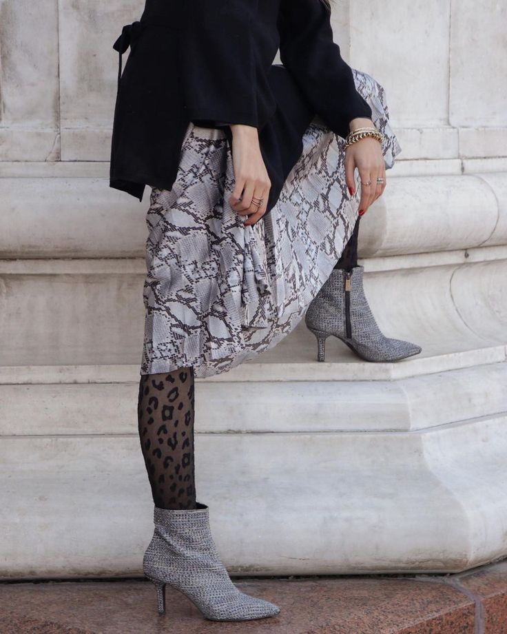 a stylish woman is posing against a stone column. she is wearing a snakeskin pleated midi skirt paired with a black sweater, leopard print tights, and sparkly heeled ankle boots.  her accessories include gold bracelets and rings. the overall style is elegant and fashionable.