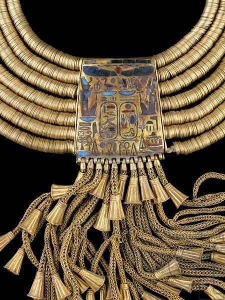 a closeup image of an elaborate gold egyptian necklace. the necklace features multiple strands of gold beads culminating in a central rectangular pendant. the pendant is adorned with colorful enamel and hieroglyphics, and from its base hangs a cascade of delicate gold tassels.