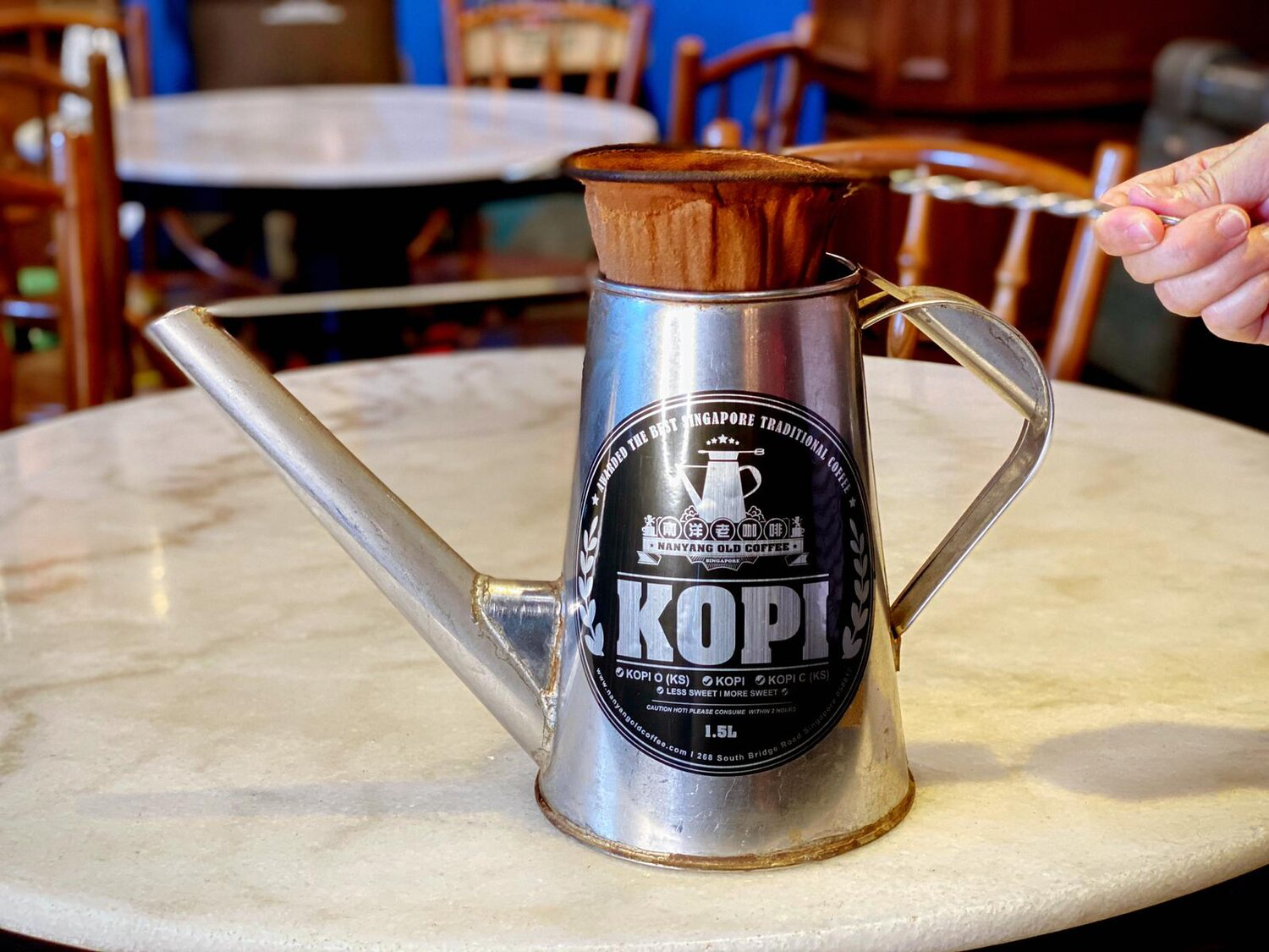 a closeup image of a silver metal coffee pot with a label that reads "nanyang old coffee" and "kopi". the pot is sitting on a marble table, with the spout facing the left side of the image. it is filled with a brown coffee filter, with a white string hanging from it.