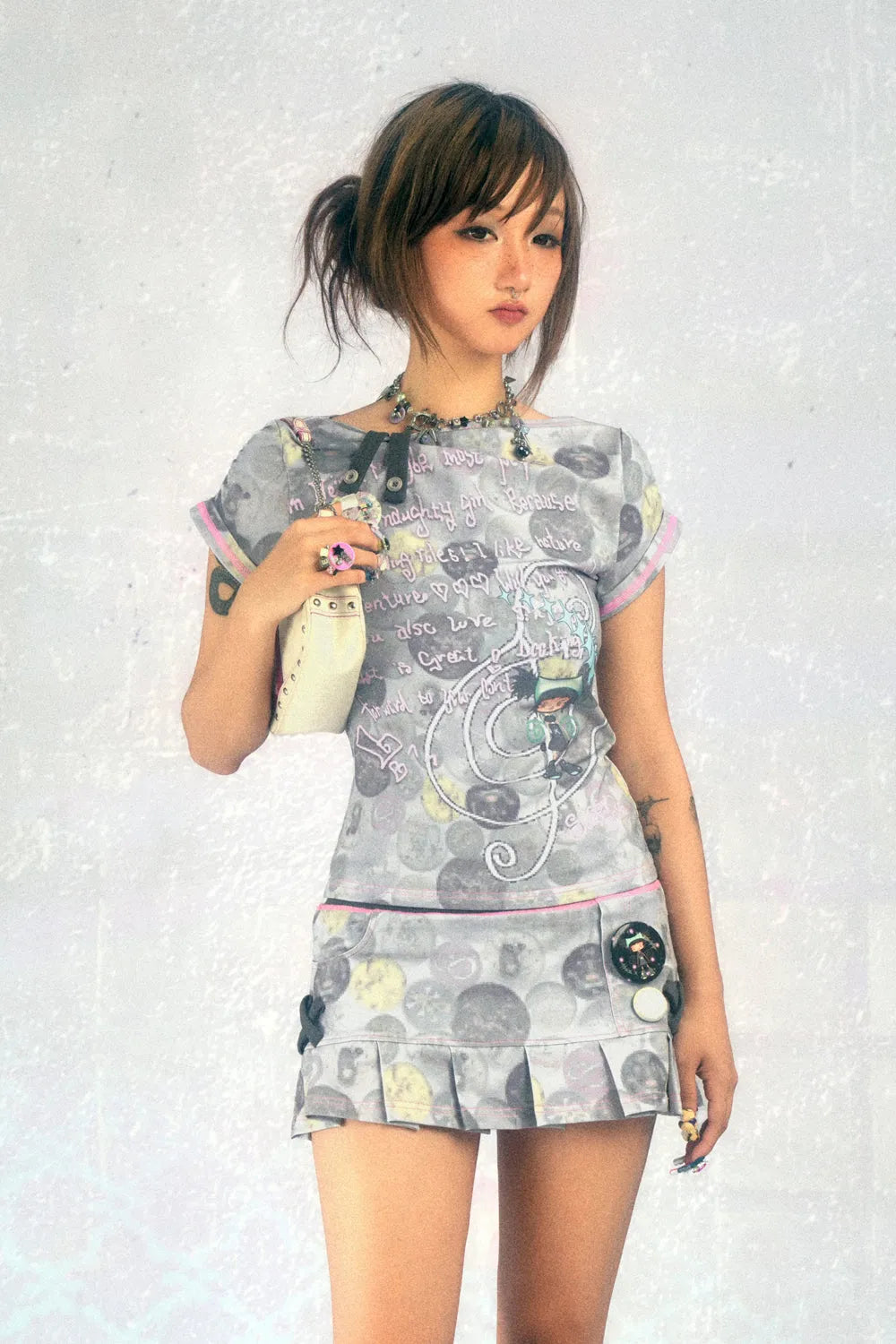 young asian woman with short brown hair is posing in a studio shot against a light grey background. she is wearing a grey pleated mini skirt and a matching grey graphic tshirt with pastel pink and light blue accents. the top features a whimsical design with text and cartoon characters.  she is holding a light beige purse and is wearing necklaces and rings. her expression is serious but not unfriendly. the overall style is casual and slightly edgy.
