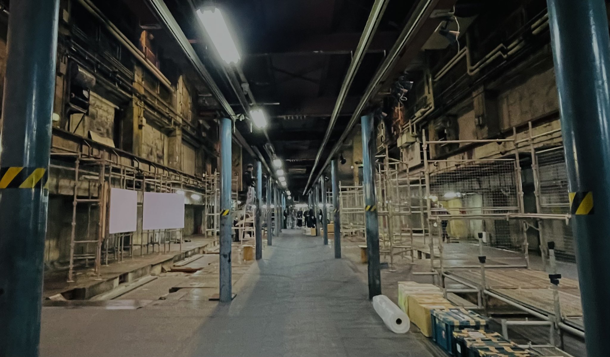 this is a photo of the interior of a large, dark, industrial building, likely an abandoned factory. there's a long central walkway flanked by heavy machinery and metal scaffolding. the walls are dark and grimy, and the lighting is dim, provided by overhead fluorescent lights. a few people are visible in the distance, emphasizing the vast scale of the space. the overall atmosphere is one of decay and abandonment.