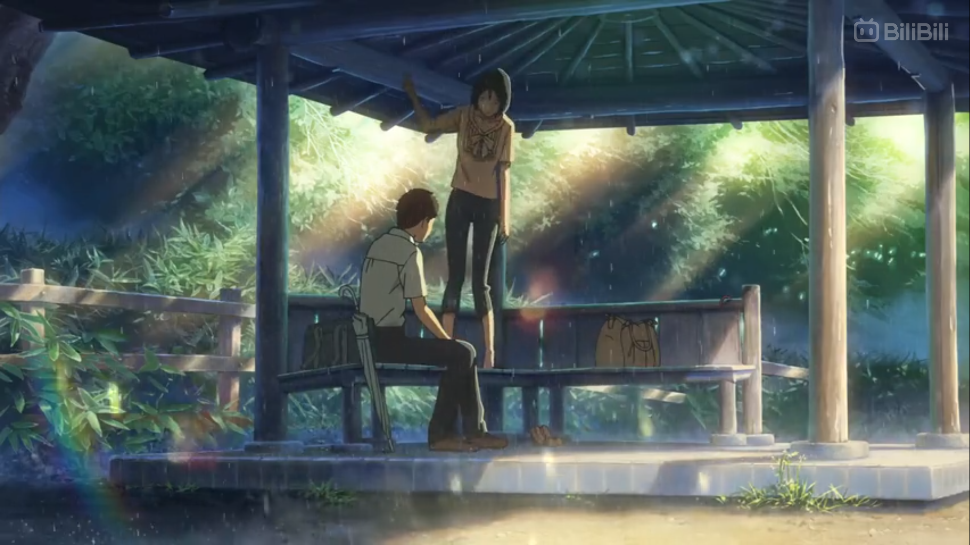 the image depicts a serene scene from an animated film. a young man and woman are taking shelter from the rain in a wooden gazebo situated within a lush, green park. sunlight filters through the trees, casting dappled light and soft shadows on the ground. the man sits on a bench while the woman stands, her hand raised as if in conversation. the overall atmosphere is peaceful and intimate. the color palette consists primarily of various shades of green and brown, reflecting the natural environment.