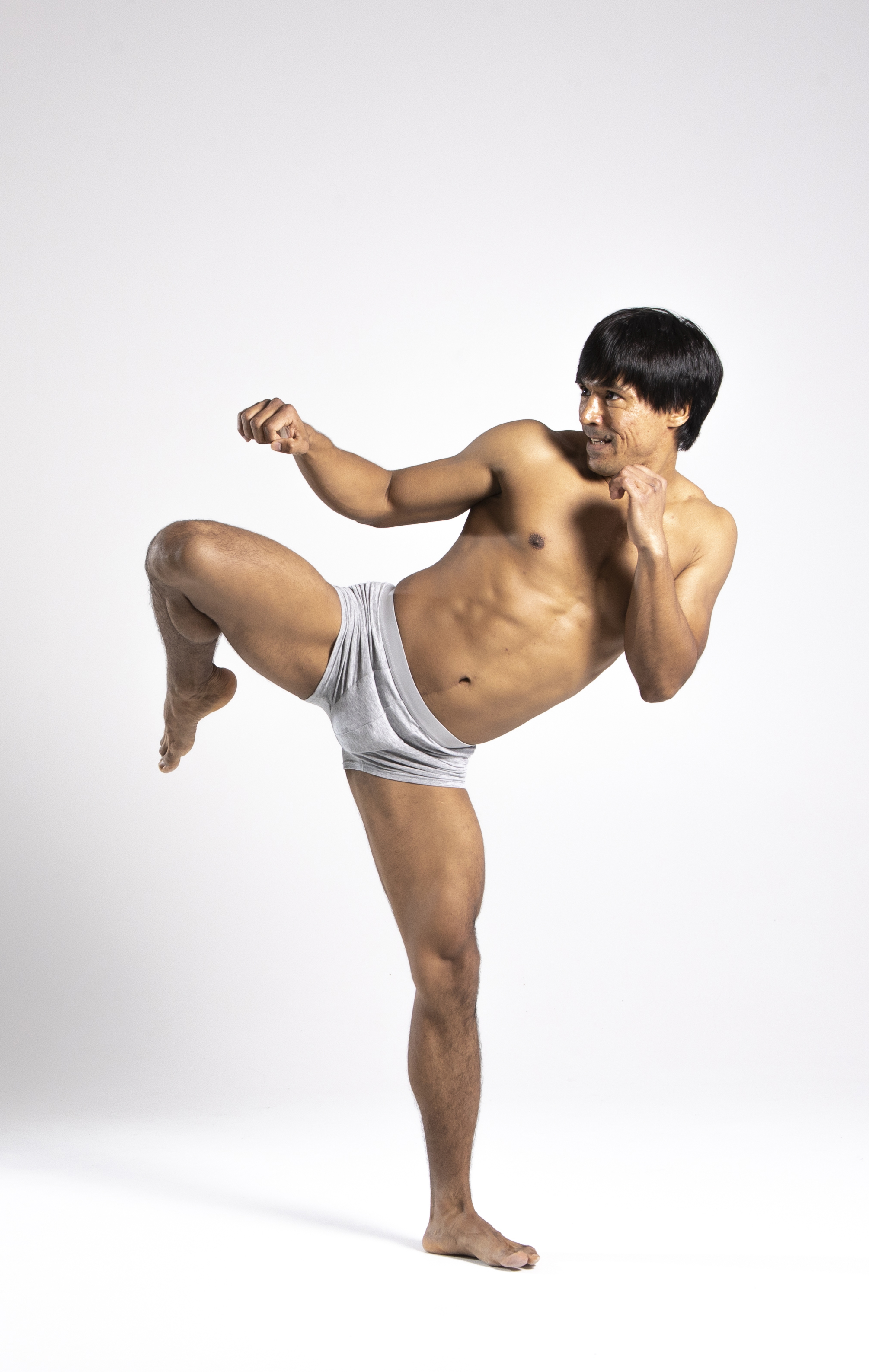 a shirtless asian man in underwear is posed in a martial arts stance against a white background. he has his left leg raised in a kick and his fists are up, ready to fight. he appears focused and strong.