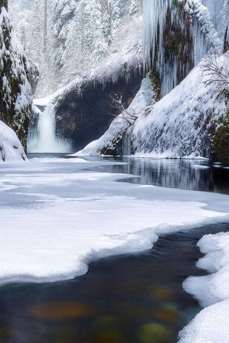 a winter landscape featuring a waterfall in the background, with snowcovered rocks and trees. icicles hang from the rock formations, and a partially frozen stream flows in the foreground, with snow and ice covering the edges.