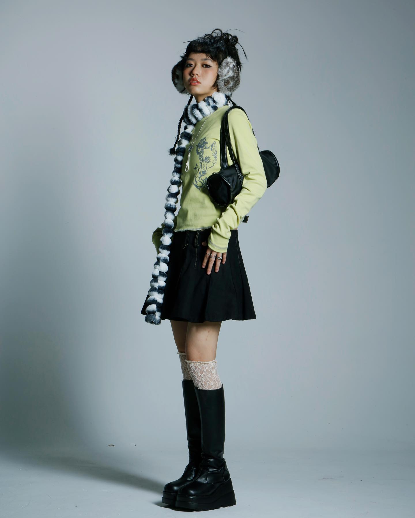 a fullbody shot of a young asian girl posing in a studio. she is wearing a lime green longsleeved shirt, a black pleated skirt, black platform boots, a black and white striped scarf, and black and white furry earmuffs. she carries a small black handbag. the background is a simple grey backdrop.  her pose is slightly angled, and she is looking directly at the camera.