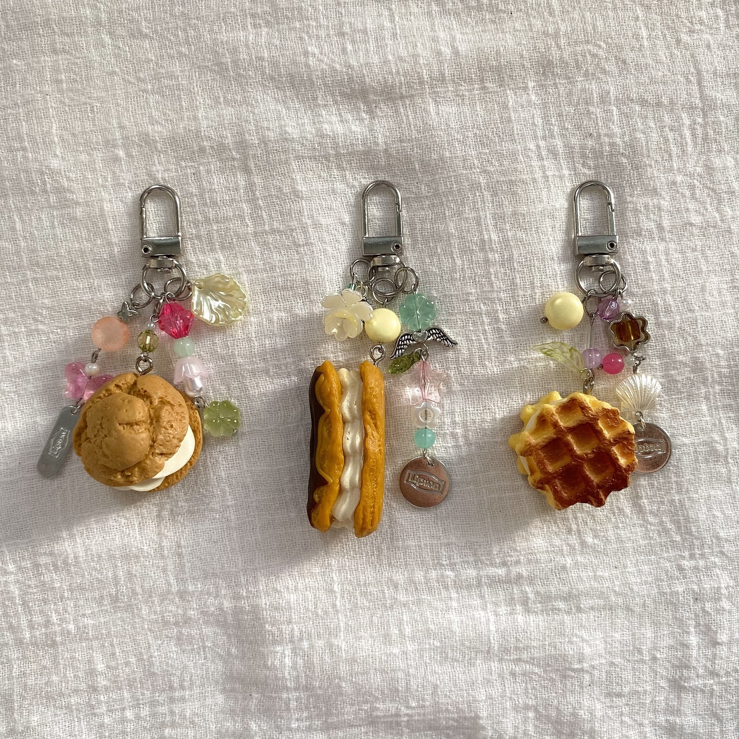 the image shows three handmade keychains with foodthemed charms. the first keychain has a cream puff charm, the second has a sandwich charm, and the third has a waffle charm. each keychain also has beads and other small charms attached. the keychains are lying on a white surface.