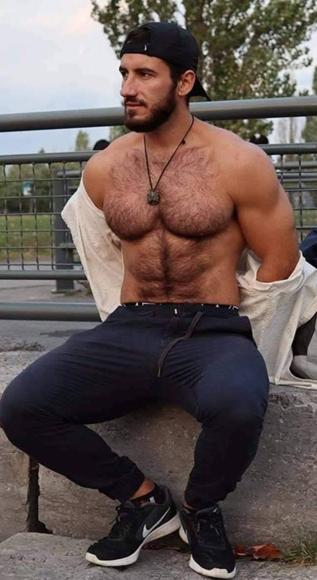 a shirtless man sits on a concrete bench, wearing black pants and a white shirt that's unbuttoned, revealing his hairy chest. he's also wearing black sneakers and a black cap. the man's gaze is to the right side of the image,  and he's posing with his hands in his lap.  there is a metal railing in the background, as well as foliage, indicating a park setting. the photo is taken during the daytime.