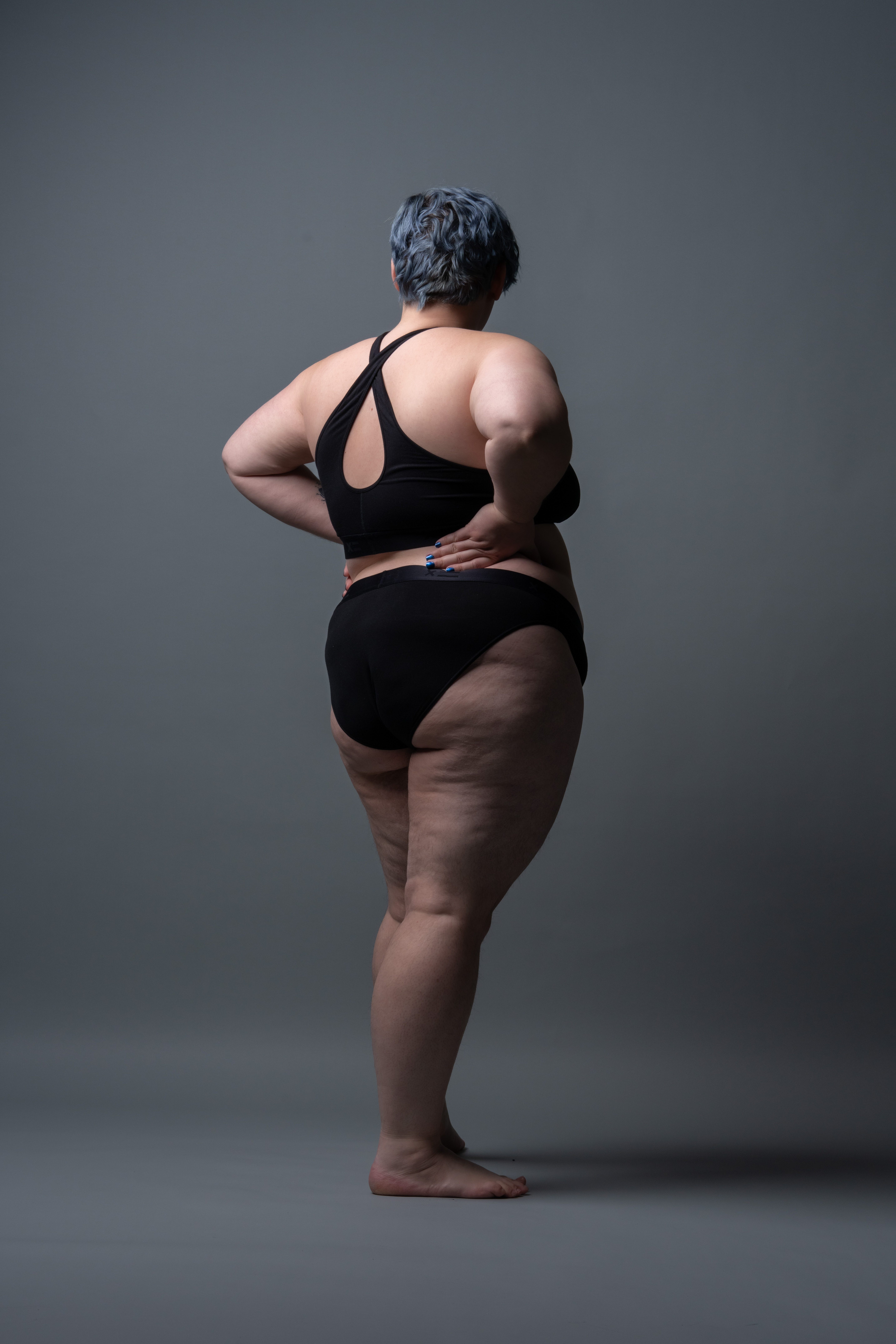 a plussize woman is seen from the back in black underwear. she is standing against a grey background with one hand on her lower back. her hair is short and blueishgrey.