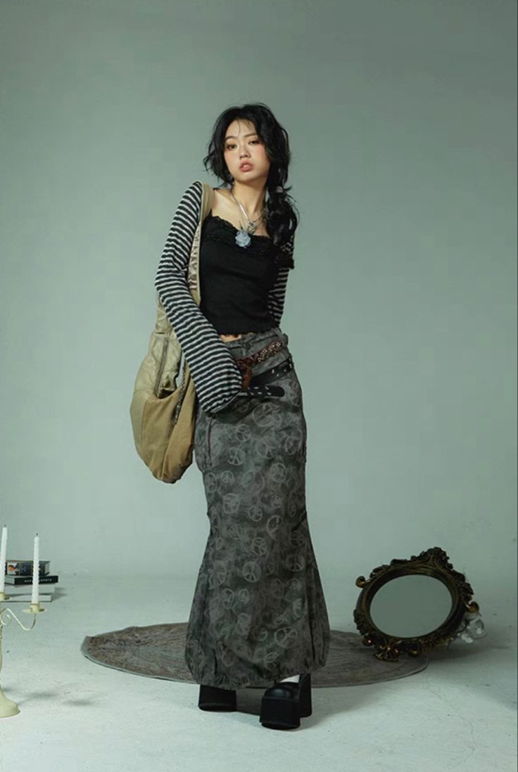 a full shot of a young woman posing in a stylish outfit. she's wearing a long, dark grey patterned skirt with a black crop top and a striped cardigan. the skirt has a flared style, and the cardigan is loosely draped over her shoulders. she's also carrying a light beige shoulder bag, and has on black platform shoes. her dark hair is styled in a way that frames her face nicely, and she's wearing a simple necklace. the backdrop is a neutraltoned studio, and a small ornate mirror rests on the floor behind her, with candles and a candlestick holder in the foreground to the left. the overall style is a mix of grunge and dark academia aesthetics.