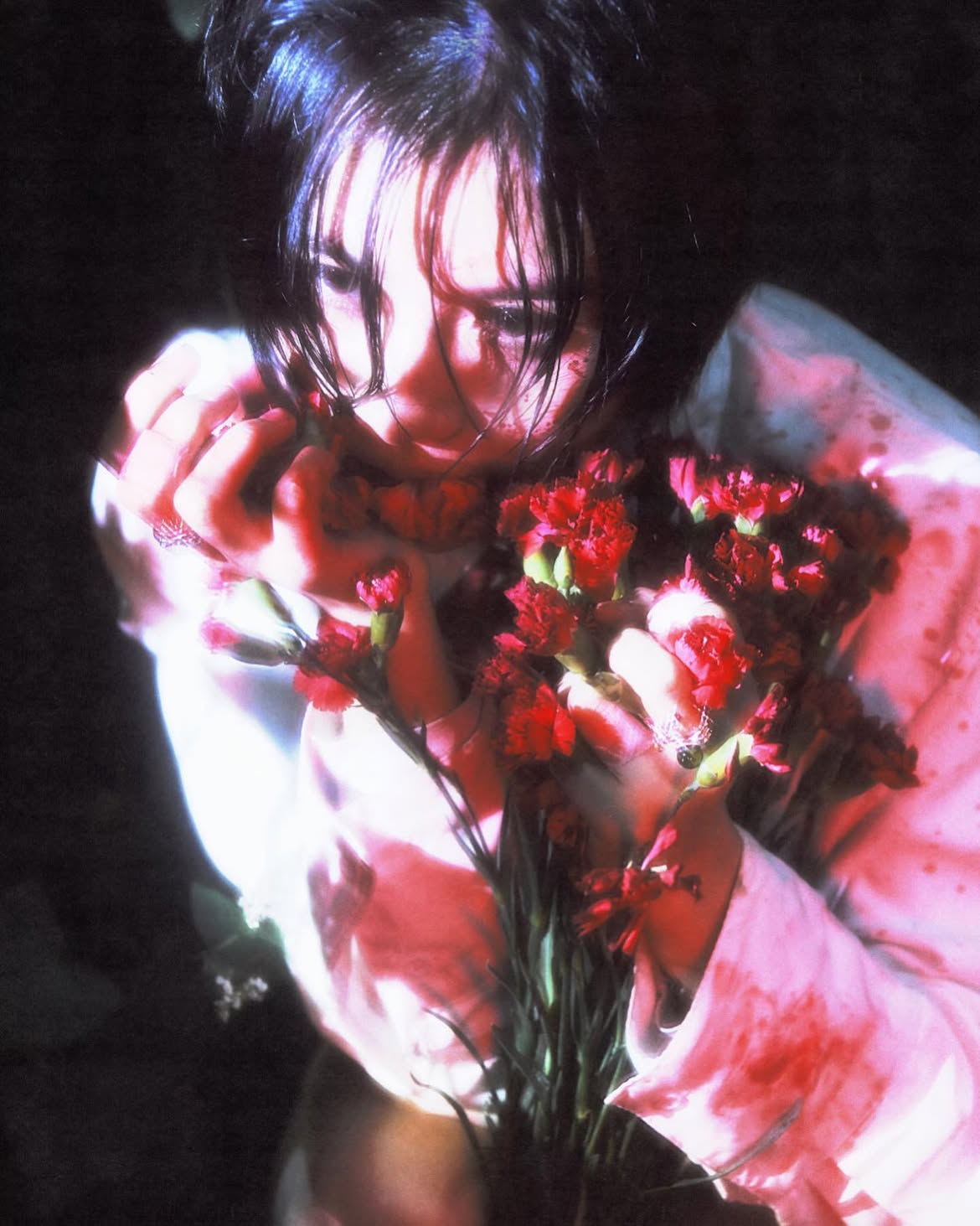 a woman with dark hair is holding a bouquet of red carnations close to her face. the lighting is moody and artistic, with low light creating a sense of emotion. her expression is intense and melancholic. the overall image has a blurred, dreamlike quality.