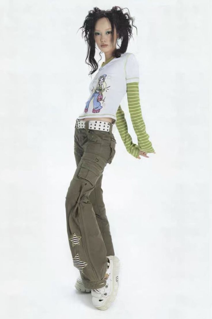 a full shot of a young woman modeling clothing. she has dark hair styled in a way popular in the early 2000s, and she wears a white longsleeved shirt with a cartoonish graphic, olivegreen cargo pants with star detailing, and white platform shoes. the background is a solid white, and she is posing casually. the overall style is reminiscent of y2k fashion.