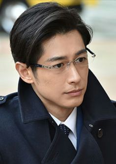 a headshot of an asian man in a suit and tie with glasses. he has short black hair and is looking slightly to the right of the camera. the background is blurred, but appears to be an outdoor setting.