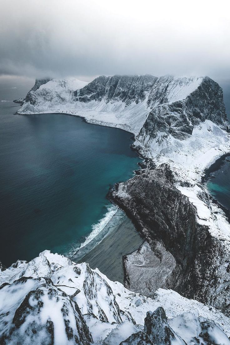 an aerial view of a rugged, snowy landscape. in the foreground, snowcovered mountain peaks stand tall. below, a strip of sandy beach meets a turquoise sea. a rocky coastline extends to a larger, snowcovered mountain island in the background, shrouded in an overcast sky.