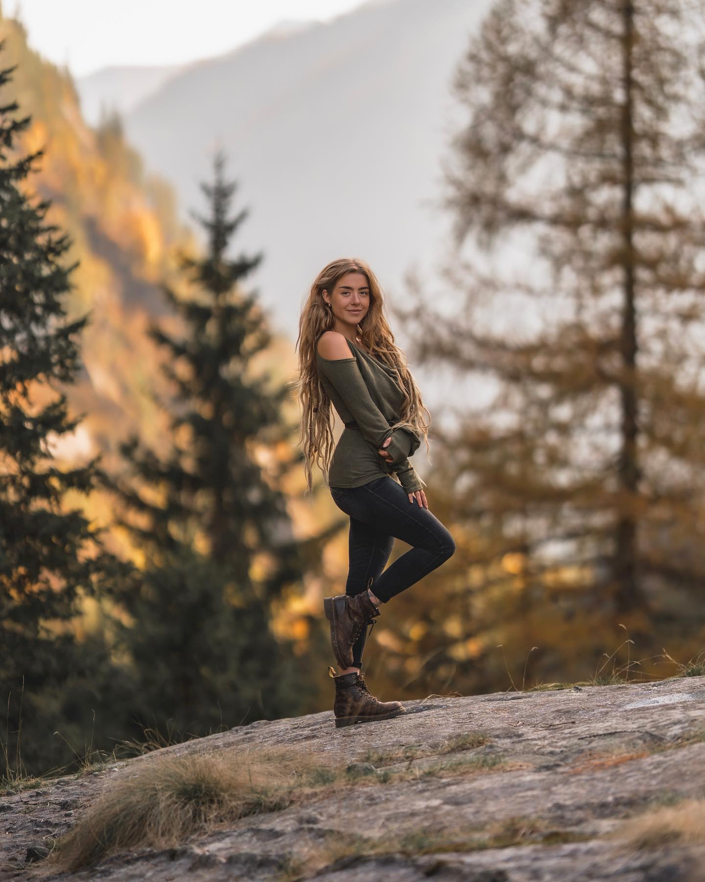 the image shows a young woman with long blonde hair standing on a rocky outcrop in a mountain forest. she's wearing an olive green offtheshoulder sweater, black leggings, and brown hiking boots. the setting is a beautiful autumnal landscape, with goldenhued trees and a soft, warm light suggesting either sunrise or sunset. the woman is positioned slightly offcenter, and she's looking directly at the camera with a gentle smile, conveying a sense of peace and serenity. the background is intentionally blurred to emphasize the woman as the main focus. the overall mood is one of calm, adventure, and connection with nature.