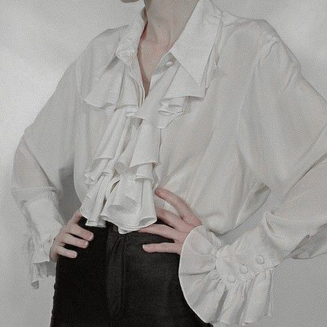 a person wearing a white, ruffled blouse with long sleeves and black pants. their hands are resting on their waist.