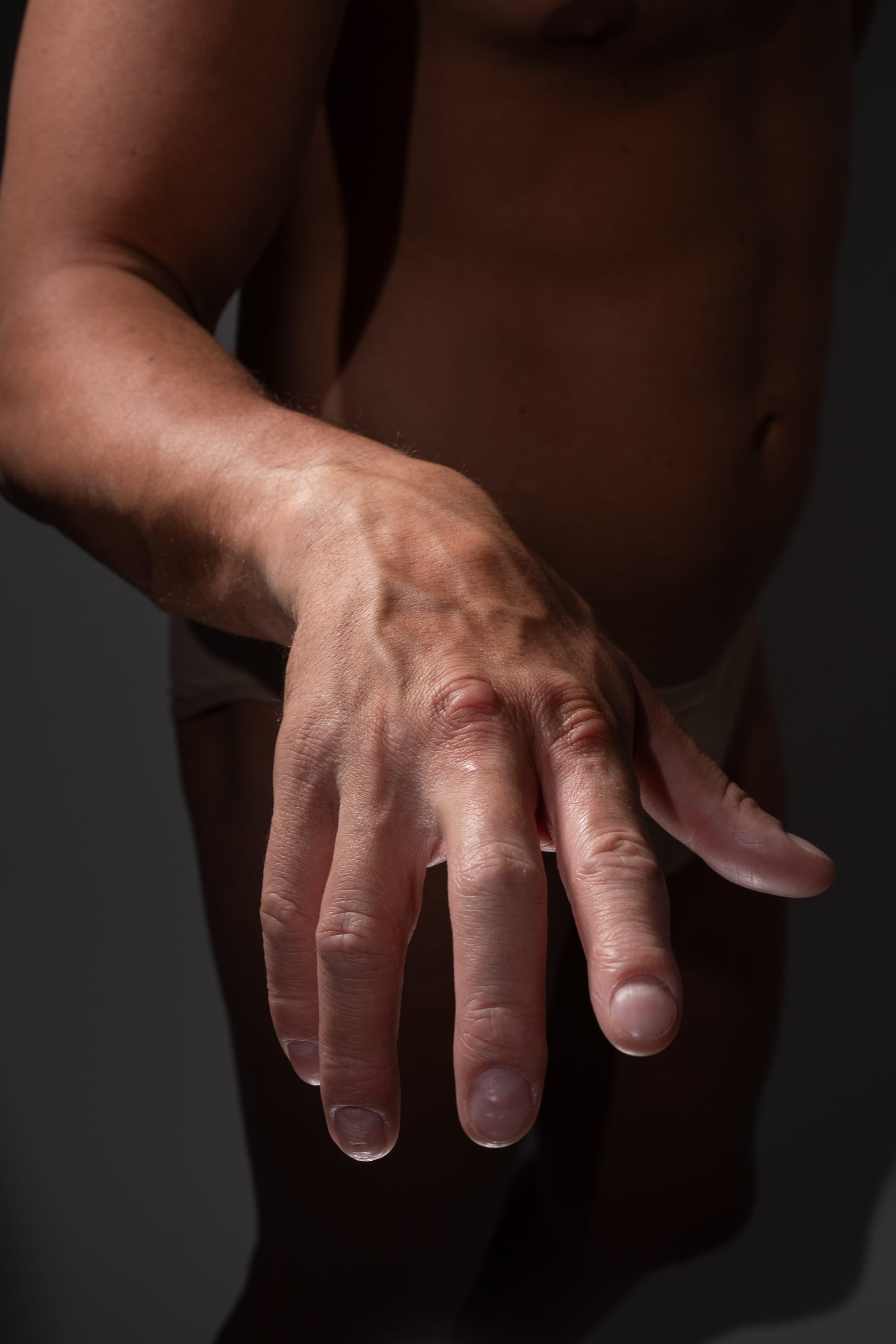 the image is a closeup of a man's arm and hand. the arm is outstretched and the hand is open. the lighting is dramatic, highlighting the contours of the muscles and veins. the background is dark and neutral, allowing the focus to remain on the hand and arm. the image has a sensual quality, emphasizing the beauty and detail of the human form.