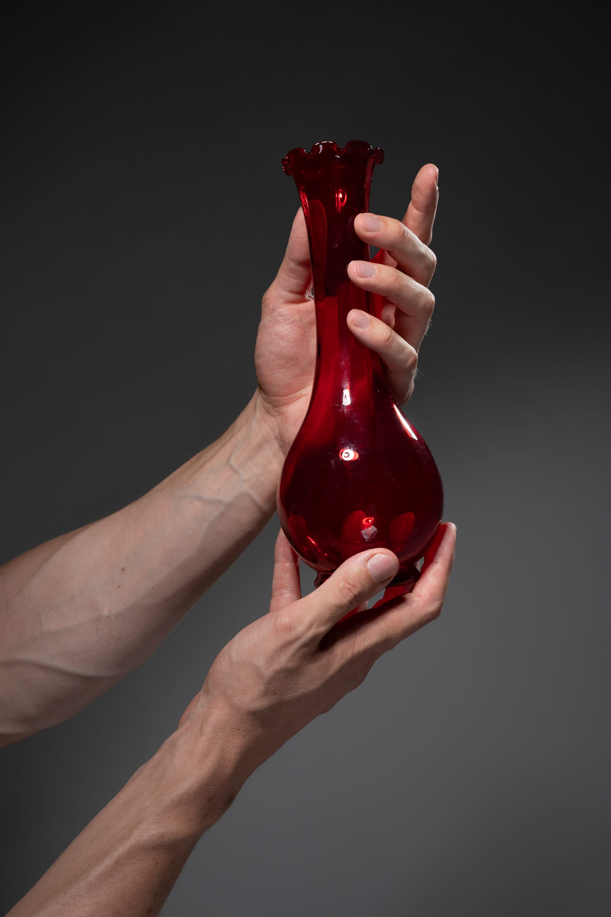 a pair of hands is carefully holding a red glass vase against a gray background. the vase has a slender neck and a bulbous base, and it reflects the light from the studio lighting.