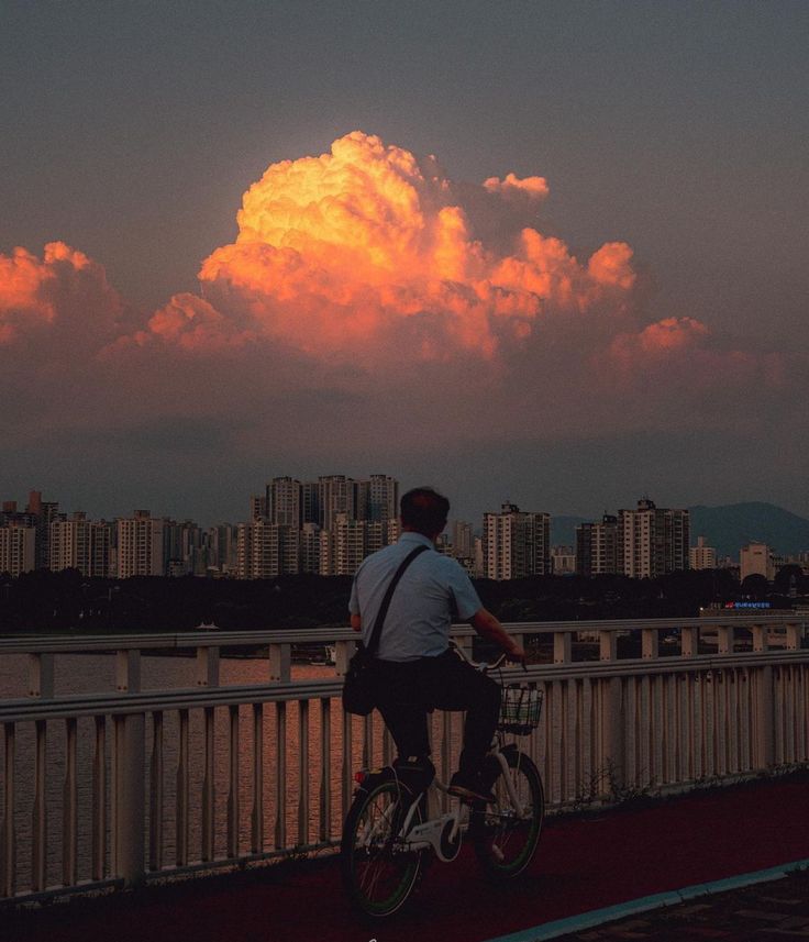 a man on a bicycle rides across a bridge overlooking a cityscape at sunset. a large, vibrant orange and yellow cloud dominates the sky above. the scene is peaceful and serene, conveying a sense of calm and solitude. the city skyline in the background is slightly hazy, adding to the moody and atmospheric quality of the image. the overall aesthetic is reminiscent of korean or east asian cityscapes.