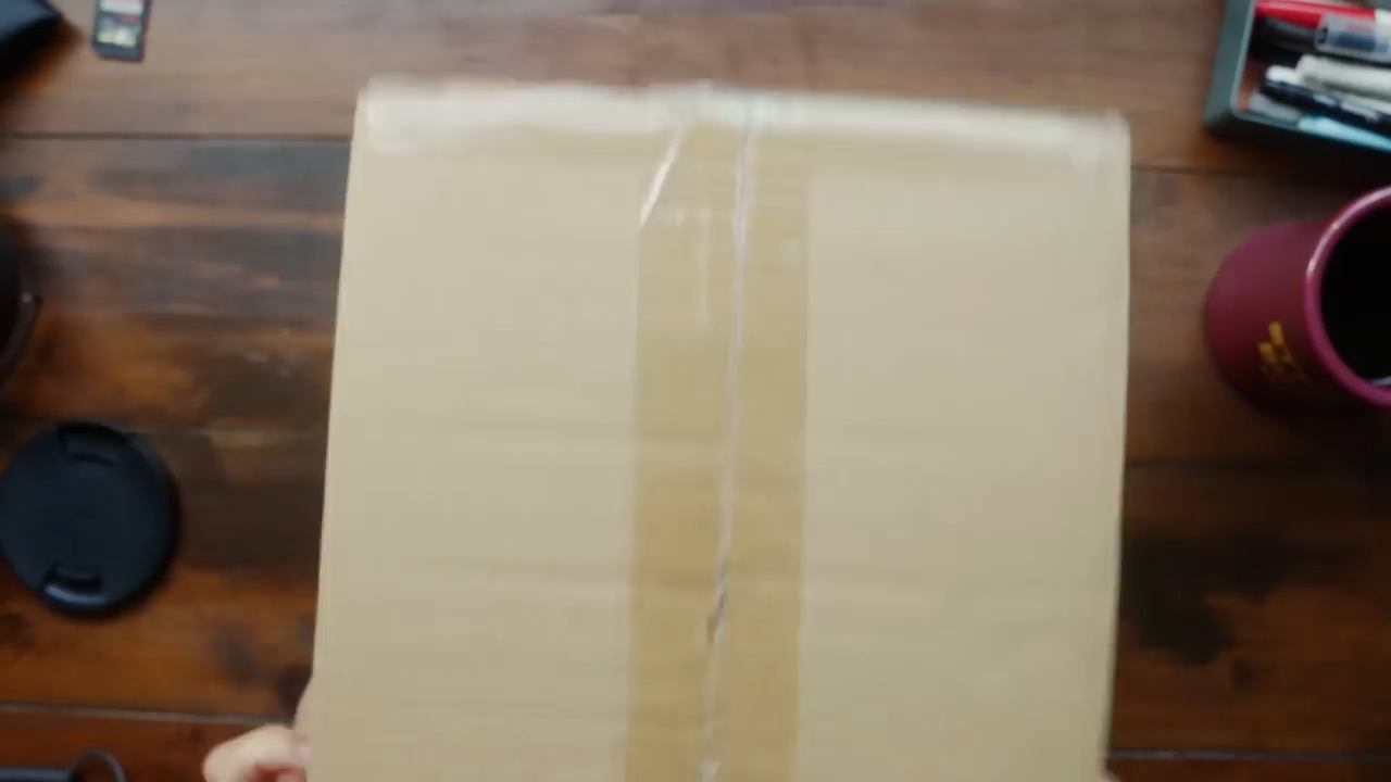 a brown cardboard box lies flat on a wooden table, with a person's hands reaching for the top to open it.  it appears to be an unboxing video or image.