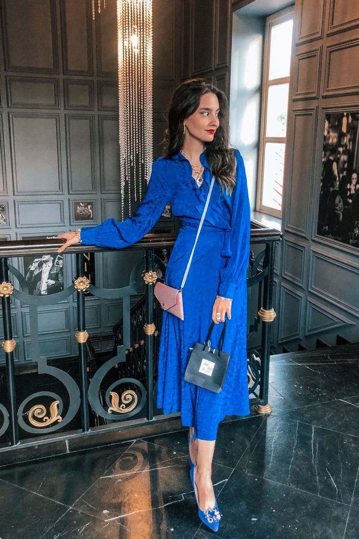 a stylish woman in a cobalt blue satin midi wrap dress stands on a marble staircase in a luxurious building. she is wearing jewelencrusted heels, a delicate necklace, and statement earrings. she carries a pale pink clutch and a small black shopping bag. the dress has long sleeves and a fitted bodice. the setting is elegant, with a dark interior and ornate railing. the overall image presents a sophisticated and stylish aesthetic.